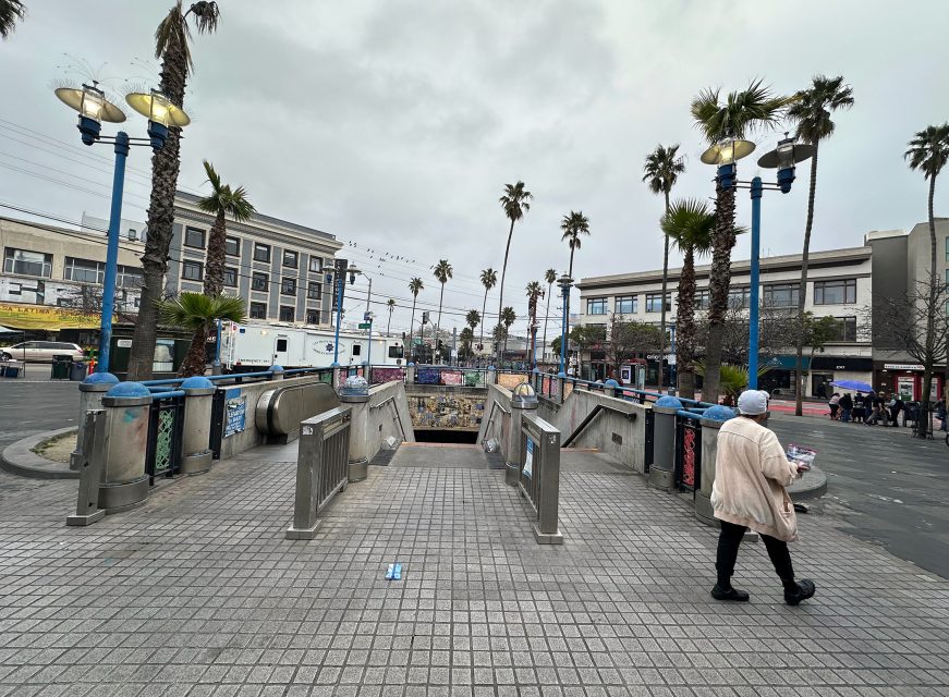 A person walks by a subway entrance in an urban plaza lined with palm trees and streetlights on a cloudy day.
