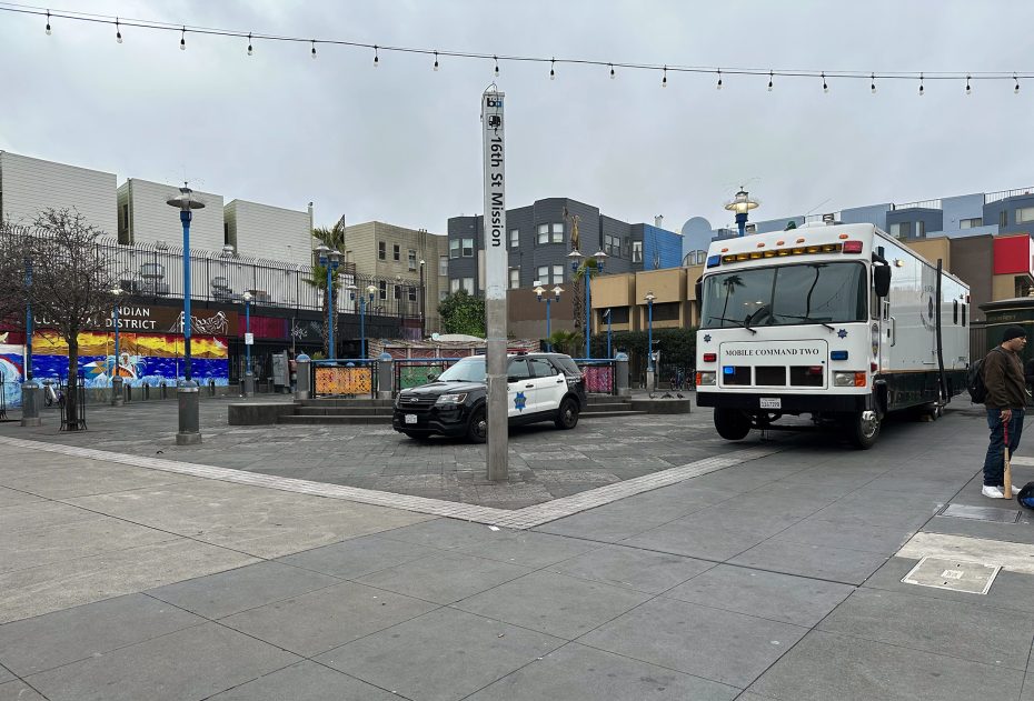 A mobile command center truck and a police car are parked in an urban plaza area surrounded by buildings.
