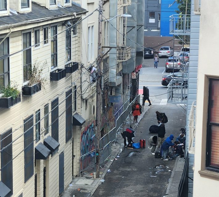 Narrow alley with people sitting and standing, surrounded by buildings with graffiti. There's a fence, utility wires, and some plants on window sills. Cars and pedestrians visible in the distance.
