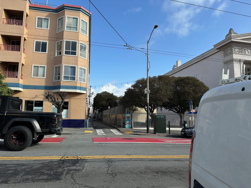 Street scene with a beige building on the left, trees lining the sidewalk, and a white van and black vehicle stopped at an intersection. Clear sky overhead.