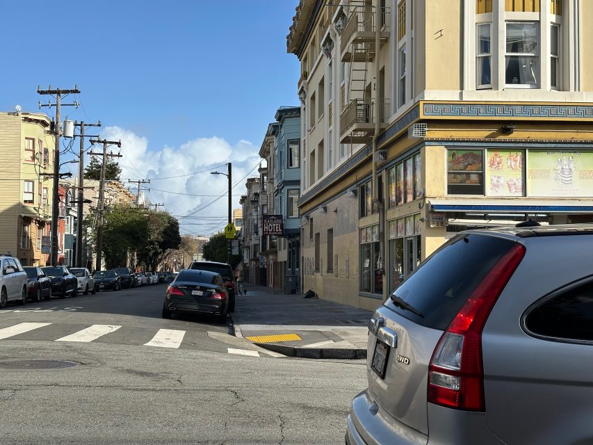 Street scene with cars parked along the curb, a visible hotel sign, and a corner building with a store. The street is lined with utility poles and leads towards distant trees under a blue sky.