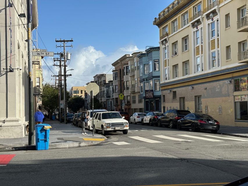 Street scene at an intersection with cars parked along the road, people walking on the sidewalk, and multi-story buildings under a partly cloudy sky.