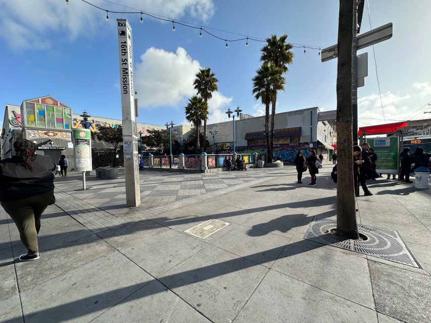 A bustling plaza with palm trees, people walking, a tall signpost, and colorful murals on building walls under a partly cloudy sky.