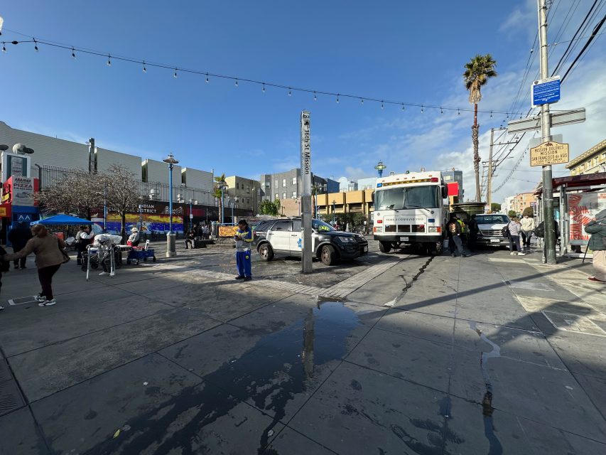 Street scene with people, parked vehicles, and a bus near a public square. Overhead string lights and a lone palm tree are visible under a clear sky.