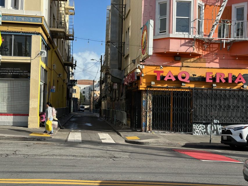 An urban street scene with a narrow alley between buildings, a closed taqueria, and people walking on the sidewalk on a sunny day.