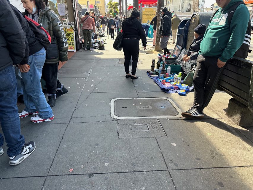 People browsing items displayed on the sidewalk. Various small goods are laid out for sale. Some individuals are standing or sitting nearby, and a mailbox is visible in the background.