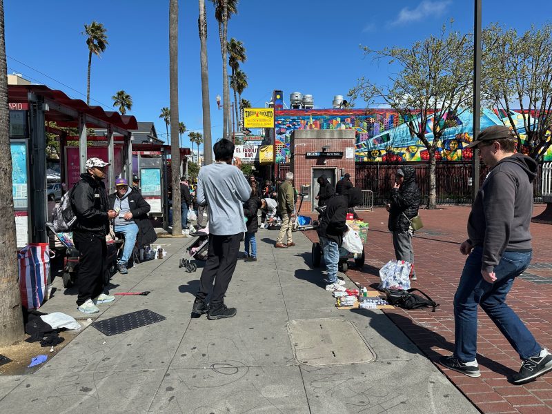 A group of people gather on a sidewalk selling and buying items near a bus stop. Palm trees and a colorful mural are in the background under a clear blue sky.