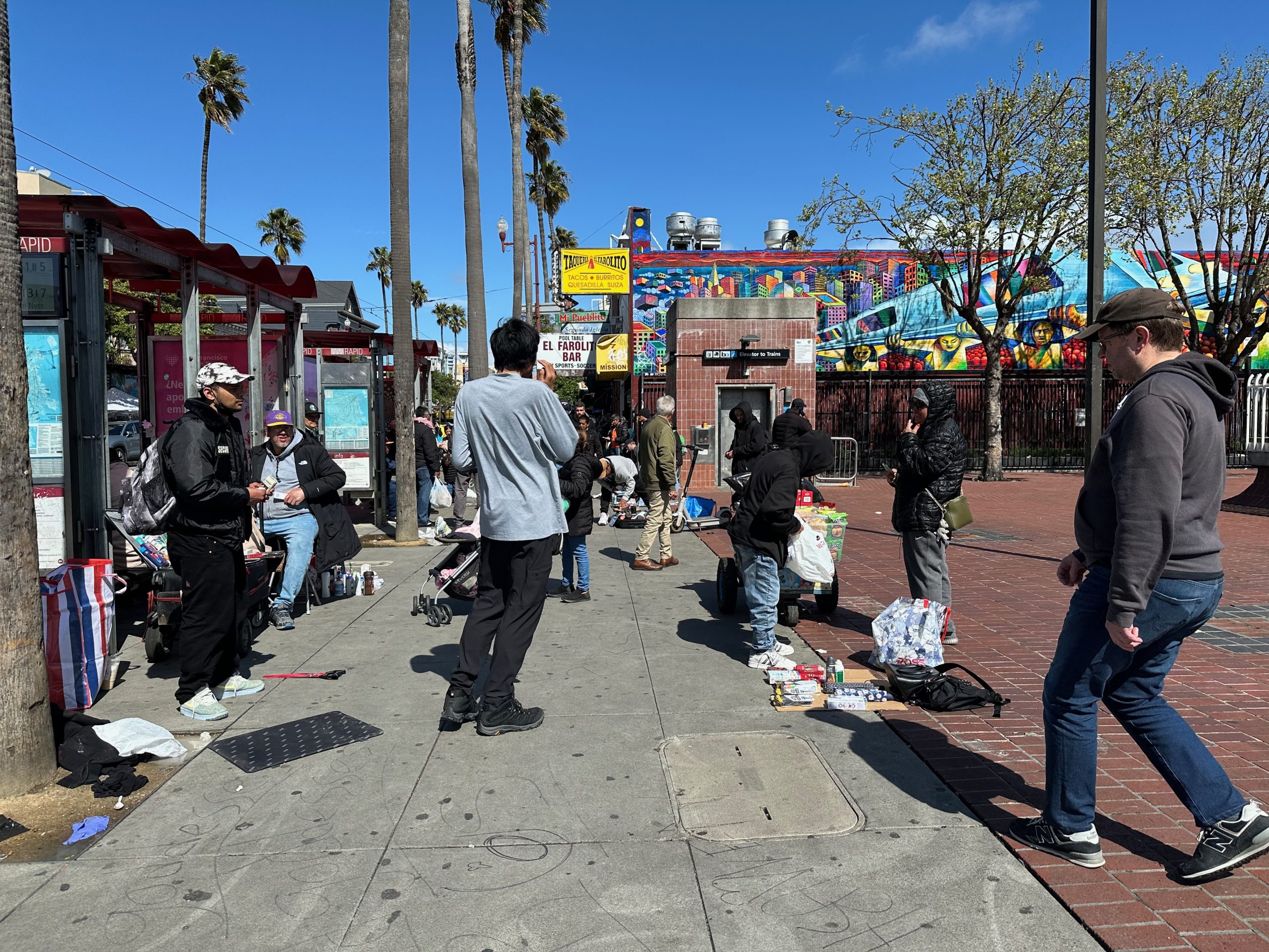 A group of people gather on a sidewalk selling and buying items near a bus stop. Palm trees and a colorful mural are in the background under a clear blue sky.