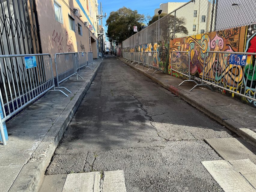 Narrow alley with graffiti-covered walls, metal barriers on both sides, and a tree at the end under a clear blue sky.