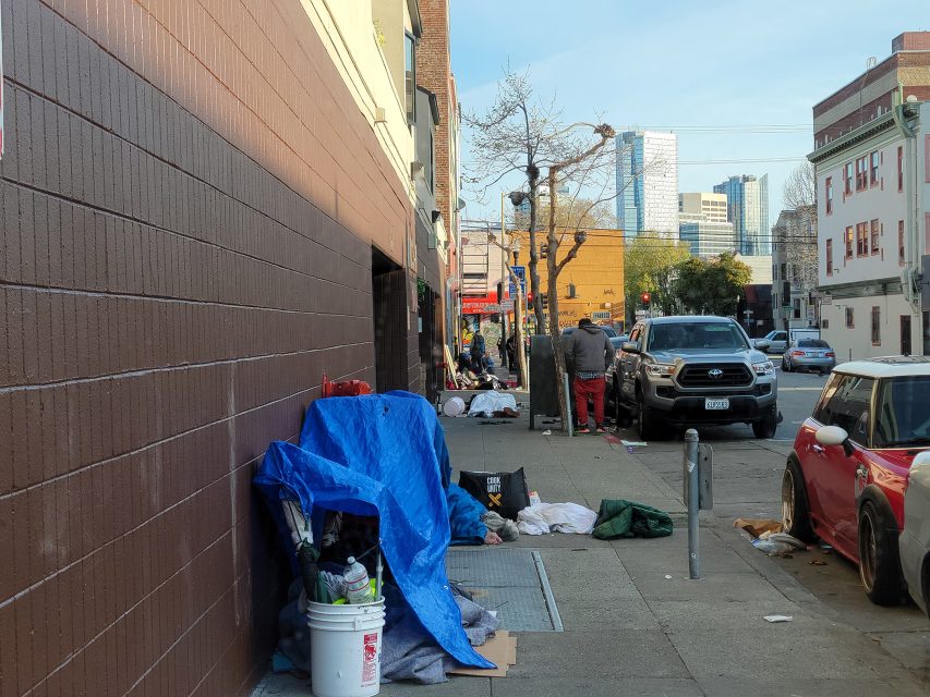 Urban street with tents and belongings along a building wall. A few people are visible in the background near parked cars. Skyscrapers are in the distance.