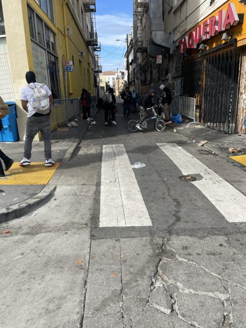 Narrow urban street scene with people standing near buildings and fences. A child rides a bicycle in the middle. The street is littered, and a taqueria is on the right.