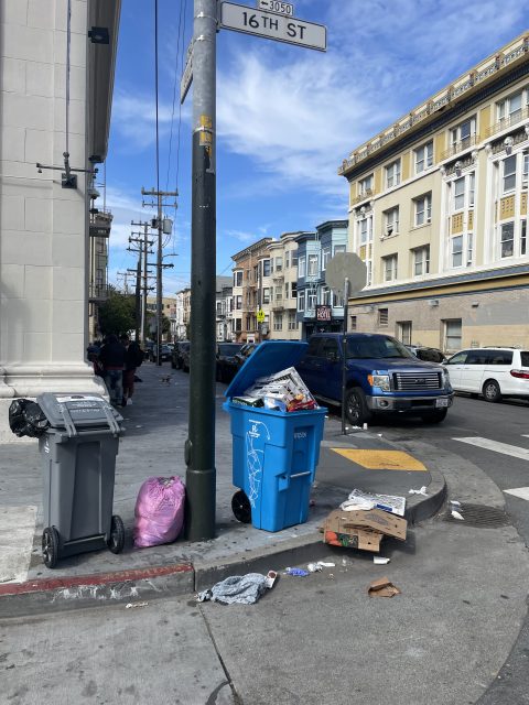Overflowing blue recycling bin and gray trash can on a city street corner. Nearby, litter and a pink bag are scattered on the sidewalk. Buildings and a parked truck are in the background.