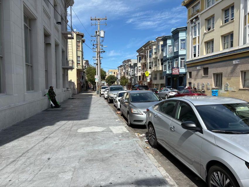 A city street with parked cars on the right side. Colorful buildings line the street under a clear blue sky. A person walks on the left sidewalk.