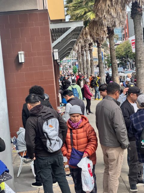 People are gathered on a sidewalk lined with palm trees, engaging in various activities. Some are standing while others are sitting near a red brick building.