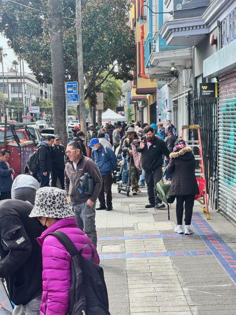 A long line of people in coats and hats stand on a city sidewalk, stretching past shops and trees.