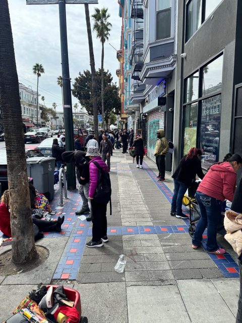 People standing in line along a city sidewalk, with various belongings and shopping carts, in front of buildings on a cloudy day.