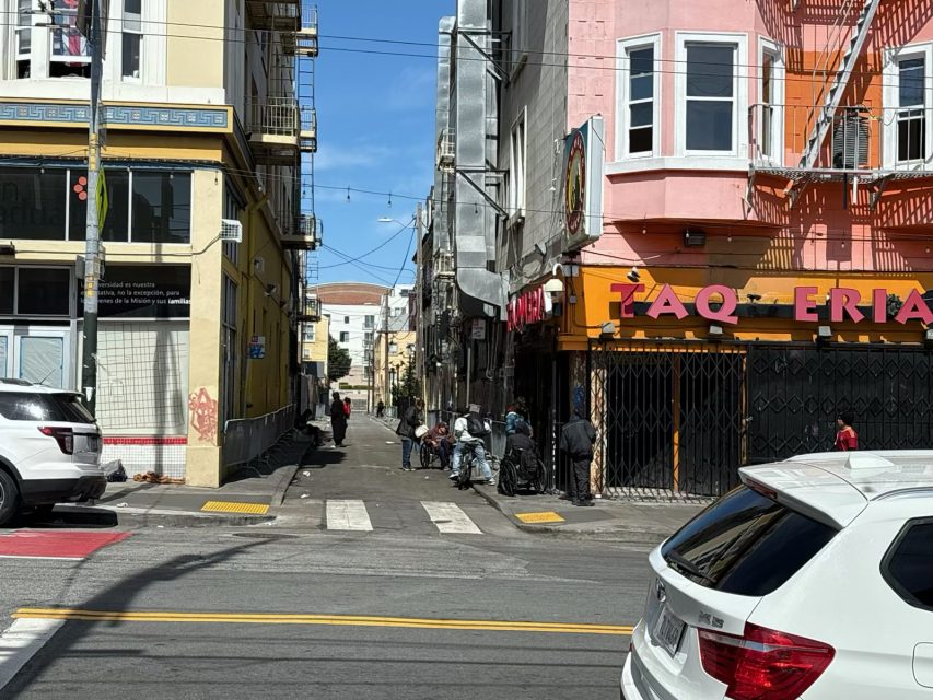 City street scene with a narrow alley, several people near a building marked "FAQ-ERIA," parked cars, and a crosswalk under a clear blue sky.