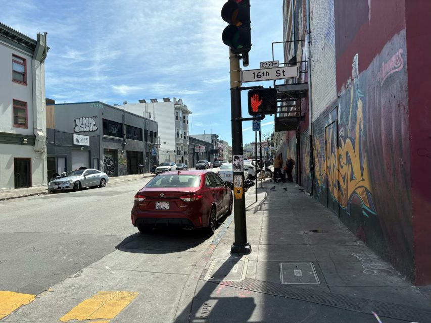 Urban street scene at 16th St with cars parked, people walking, and a visible graffiti-covered wall. A 'don't walk' signal is illuminated at an intersection under a clear sky.