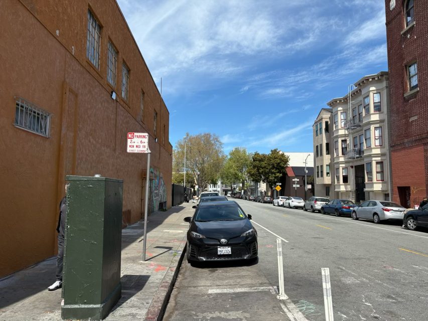 A black car is parked on a street next to a building under a blue sky. Adjacent buildings line the street with parked cars. A "No Parking" sign is visible.
