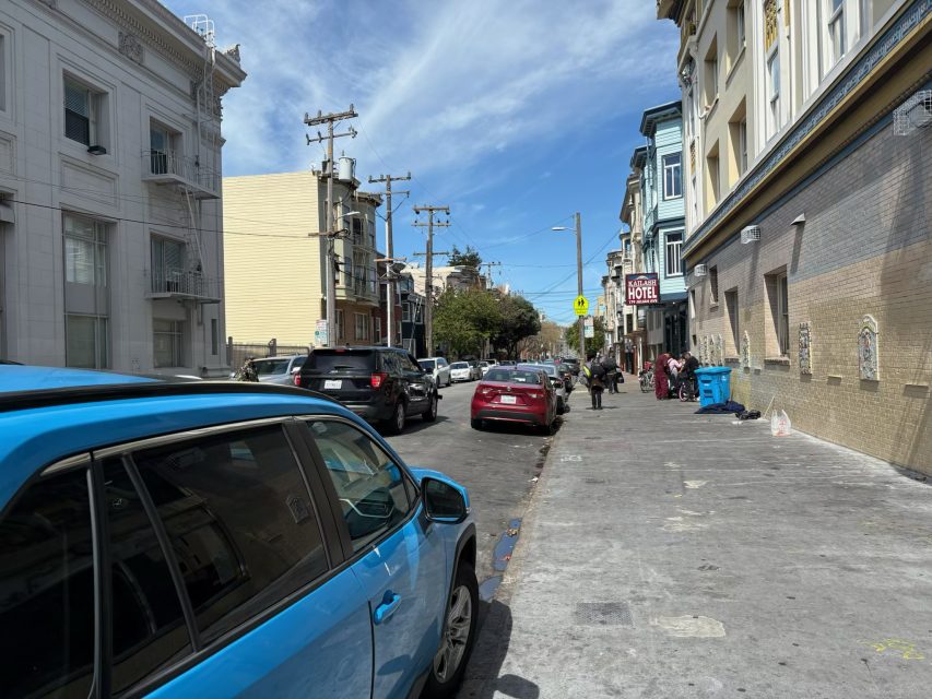 Street view with parked and passing cars on a sunny day. Buildings line both sides, and a few people are walking on the sidewalk.