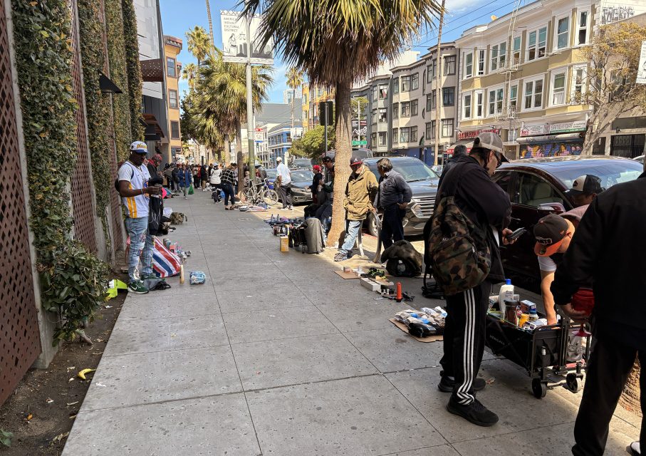 People browsing items displayed on the sidewalk of a busy street with palm trees and parked cars. Buildings line the opposite side of the street.