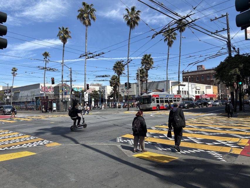 An urban intersection with people crossing, a person on a scooter, and palm trees lining the street. A bus and various buildings are visible in the background.