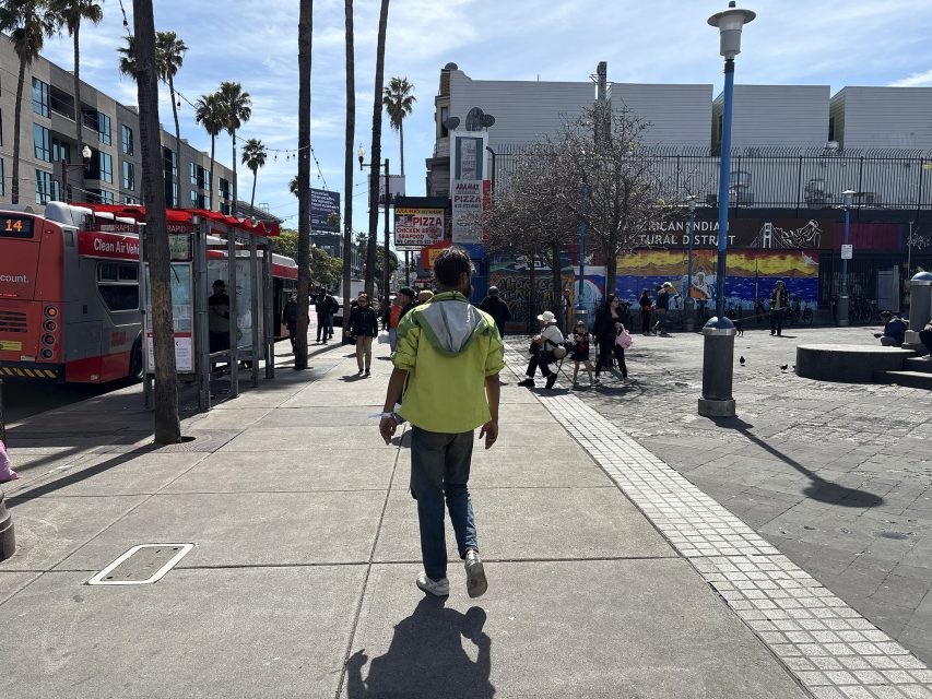 Person in a green jacket walks on a sunny street with palm trees, passing a parked bus and several people. Urban setting with buildings and murals in the background.