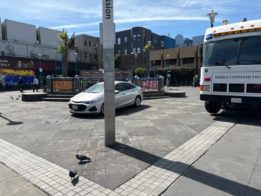 City plaza scene with a white car, several pigeons on the ground, and a mobile command truck on the right. Background includes modern buildings and a mural on the fence.