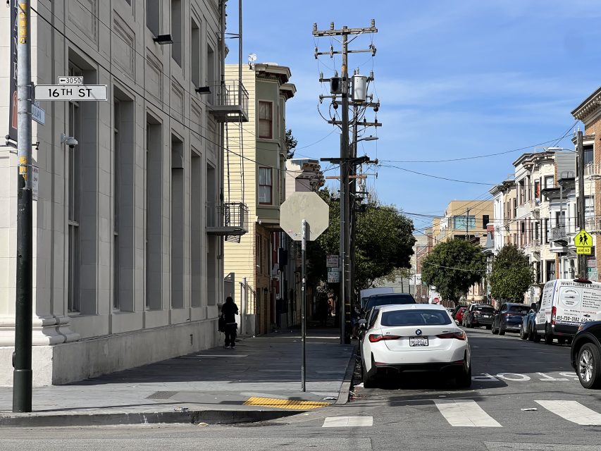 Urban street scene at 16th St. Plaza with a white car parked next to a stop sign. Tall buildings line the street under a clear sky, and a pedestrian strolls along the bustling sidewalk.