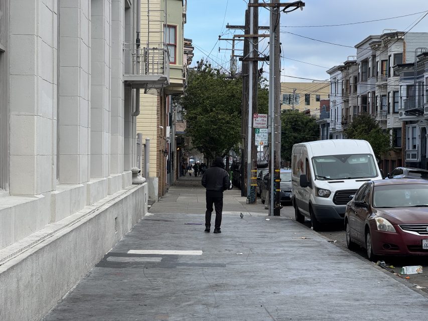 A person walks down a city sidewalk lined with buildings, parked cars, and utility poles. A white van and a red car are parked on the street. Trees are visible in the background.