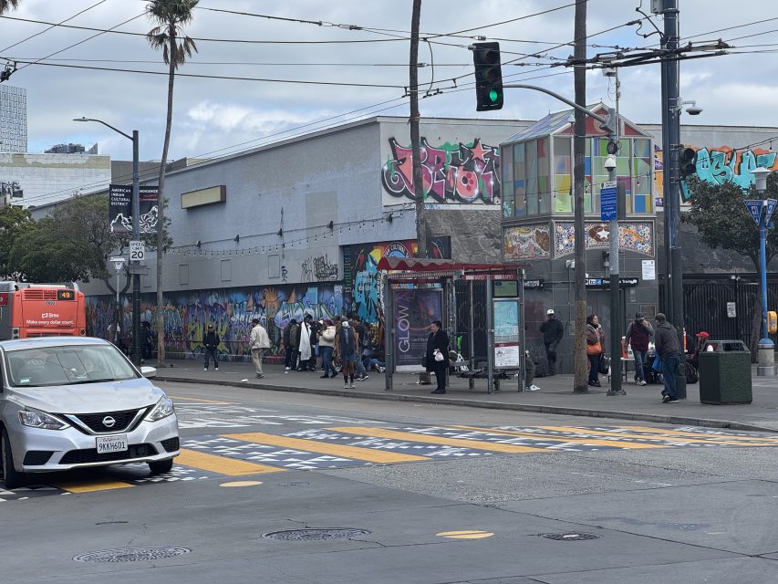 Urban street scene with a busy intersection, colorful graffiti on building walls, pedestrians waiting at a bus stop, and a white car in the foreground.