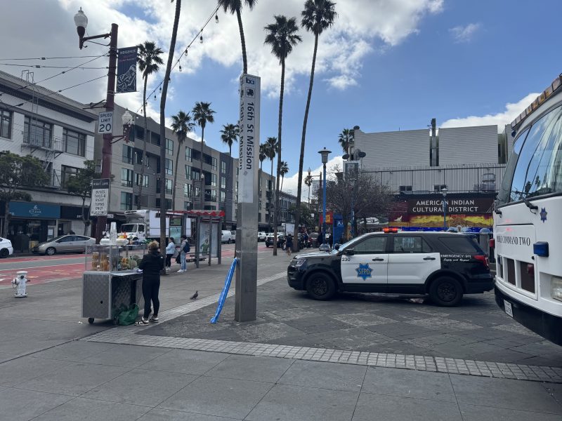 Street scene at 16th St Mission; vendor cart, pedestrians, police cars, bus, and palm trees. American Indian Cultural District sign in the background.