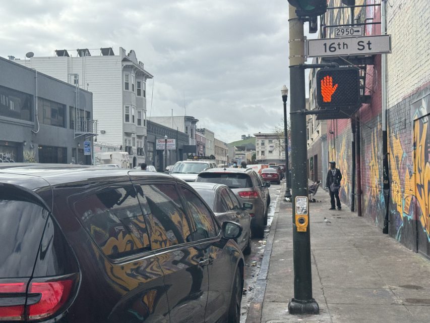 A street view with parked cars, a pedestrian walking on the sidewalk, and a street sign for 16th St. The pedestrian signal shows a red hand. Buildings are visible in the background.