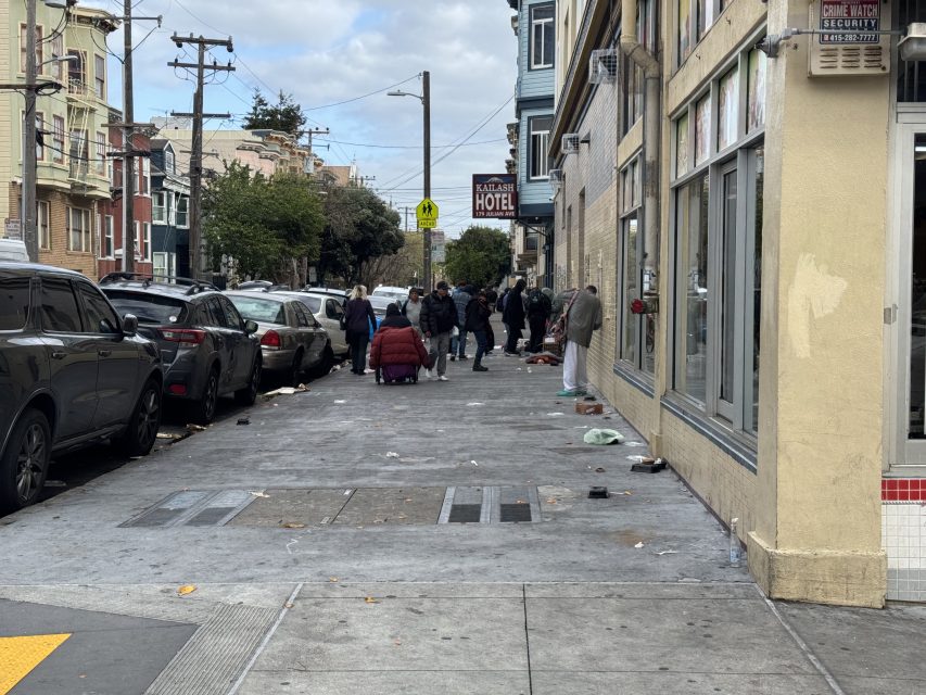 A city street with a group of people gathered on the sidewalk. Nearby are parked cars, buildings, and a "Hotel" sign. Various items are scattered on the ground.