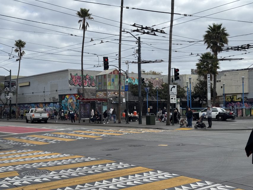 Street scene with people at a bus stop, colorful graffiti on buildings, a crosswalk with geometric patterns, and palm trees under a cloudy sky. Traffic signals and power lines are visible.