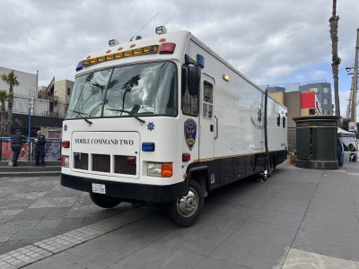 Mobile command vehicle parked on a city street near a sidewalk. Buildings and a tree are visible in the background under a cloudy sky.