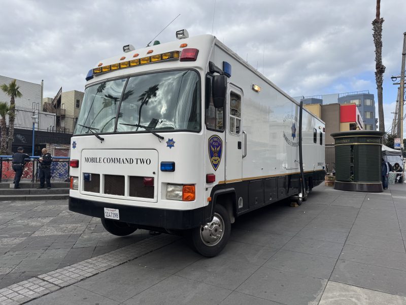Mobile command vehicle parked on a city street near a sidewalk. Buildings and a tree are visible in the background under a cloudy sky.