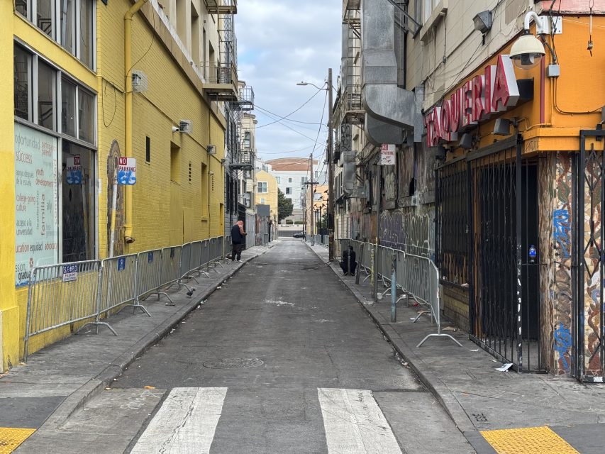 Empty urban alley with metal barricades on both sides, yellow and orange buildings, a taqueria, and a person in the distance. Overcast sky.