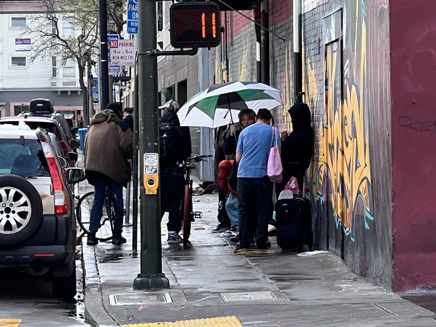 People standing on a wet city sidewalk at 16th St. plaza, some with umbrellas. A person films with a camera as vibrant graffiti and parked cars line the street.