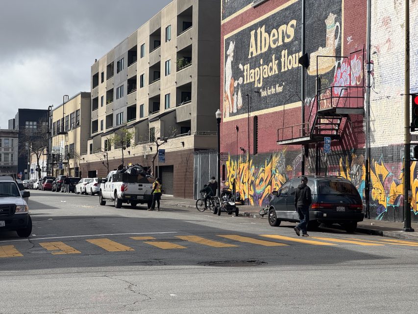 Urban street scene with a mural on a building, parked cars, and people walking and cycling. Crosswalk and graffiti are visible. Overcast sky.