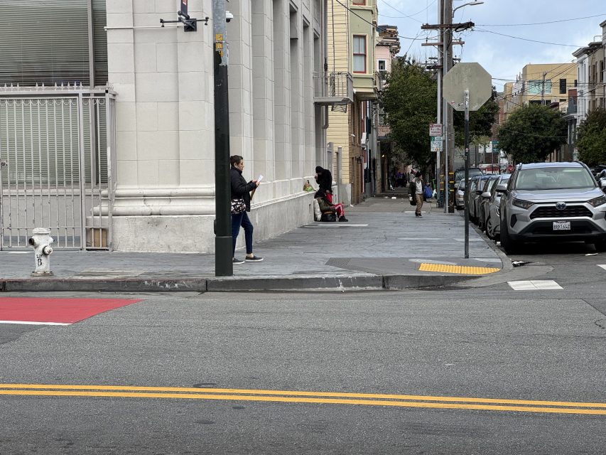 A woman stands on a city sidewalk using her phone near a corner with a fire hydrant. A person sits on a ledge in the background, and several cars are parked on the street.