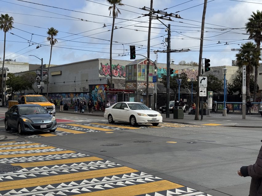 Street intersection with cars, a school bus, and colorful graffiti-covered buildings. Pedestrians cross at a marked crosswalk. Palm trees line the street, and traffic lights are visible.