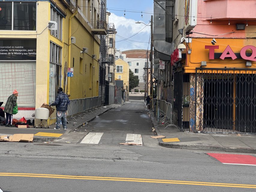 Urban alleyway with trash scattered on the ground. Two people stand near a trash bin on the left. Buildings line the alley, including one with a "TAQU" sign on the right.