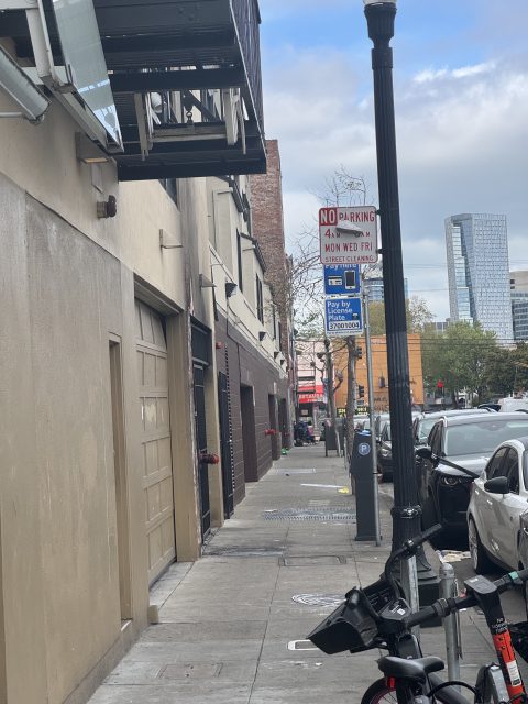 Urban street with parked cars, bicycles, a no parking sign, and buildings lining the sidewalk under a cloudy sky.
