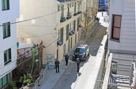 Police officers and a patrol car are situated in an urban alleyway, with graffiti on the walls and a few pedestrians in the distance.