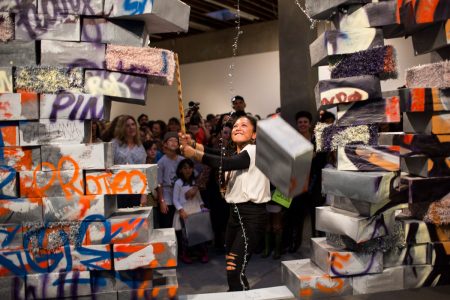 A person swings a tool to break through a wall made of graffiti-covered blocks, while a crowd watches in a gallery setting.