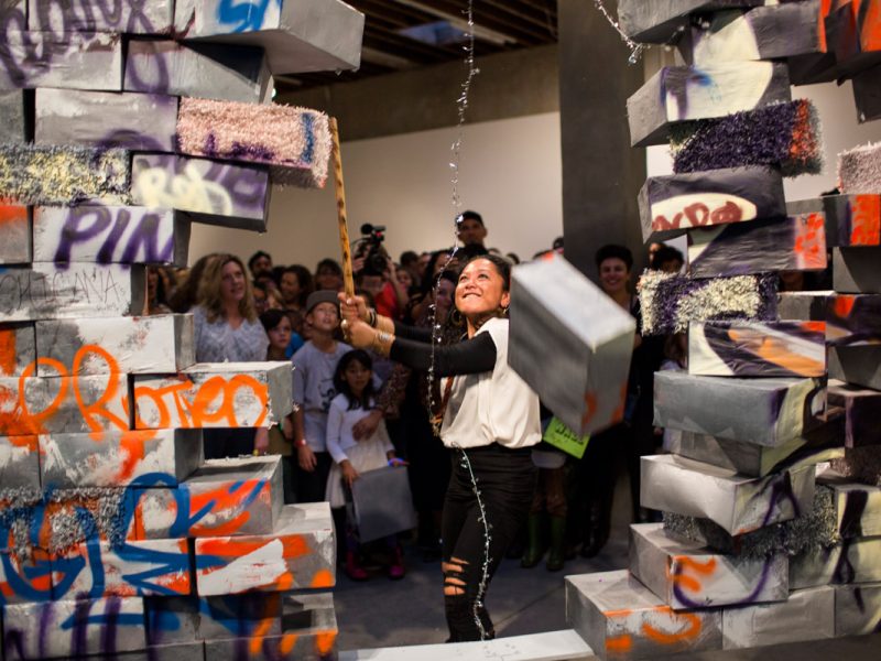 A person swings a tool to break through a wall made of graffiti-covered blocks, while a crowd watches in a gallery setting.