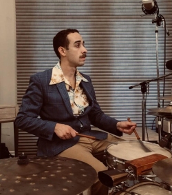 A person in a blue suit and patterned shirt plays the drums in a room with closed blinds.
