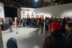 A group of people in a gallery space listens to a speaker at a microphone, surrounded by displayed artwork on the walls.