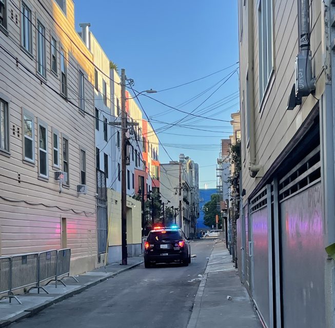 A police car with flashing lights is parked in a narrow urban alley near the lively 16th Street BART plaza, surrounded by towering buildings.